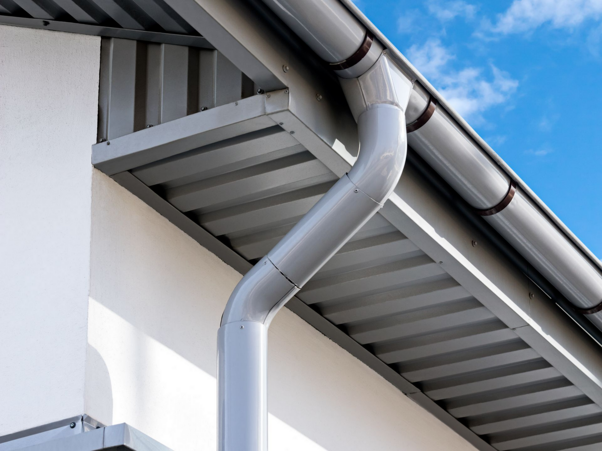 Gouttière et descente pluviale grises sur un bâtiment blanc au toit en tôle ondulée, sur fond de ciel bleu.