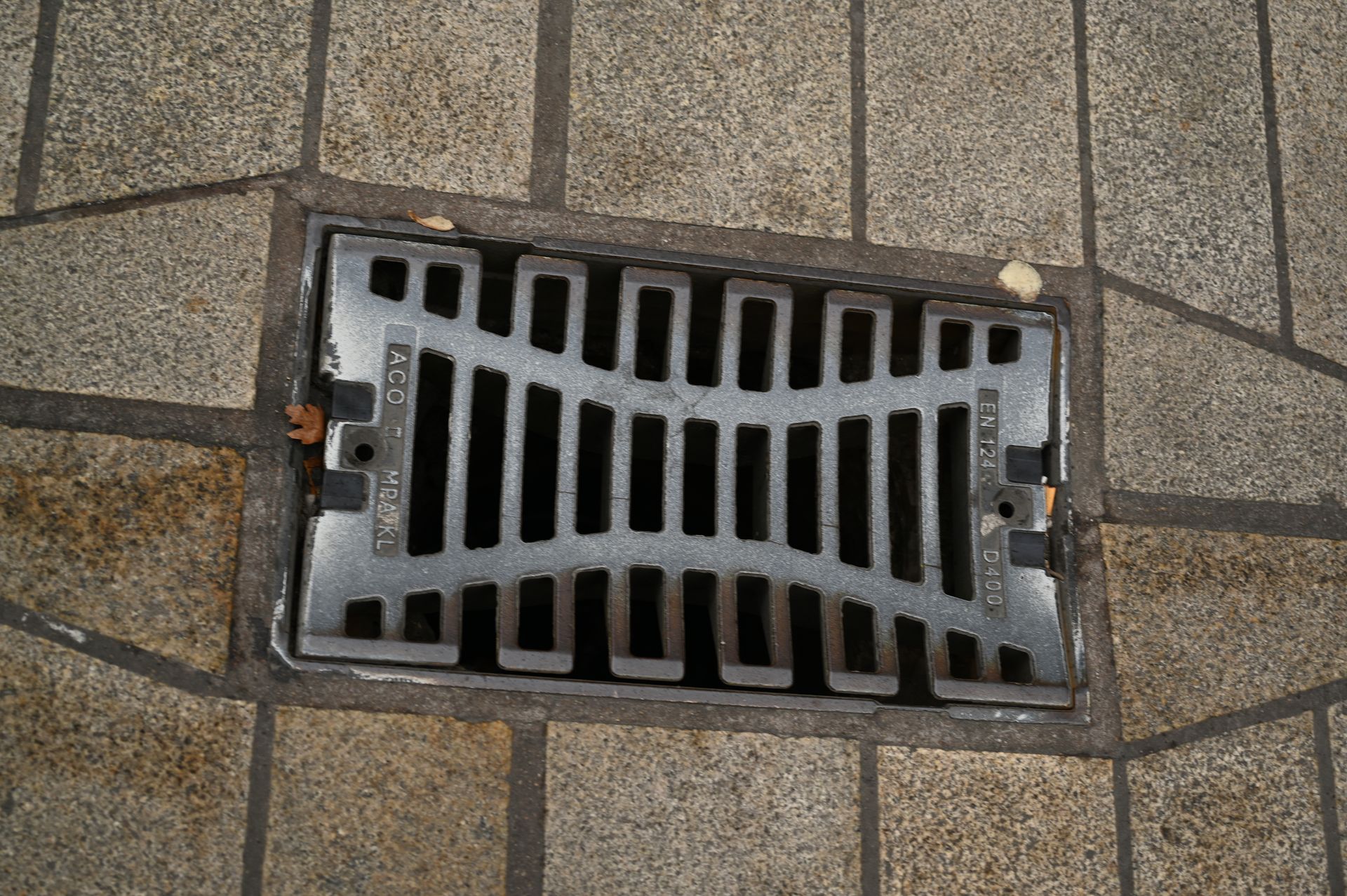Plaque d'égout métallique sur trottoir, avec un motif de barres rectangulaires.