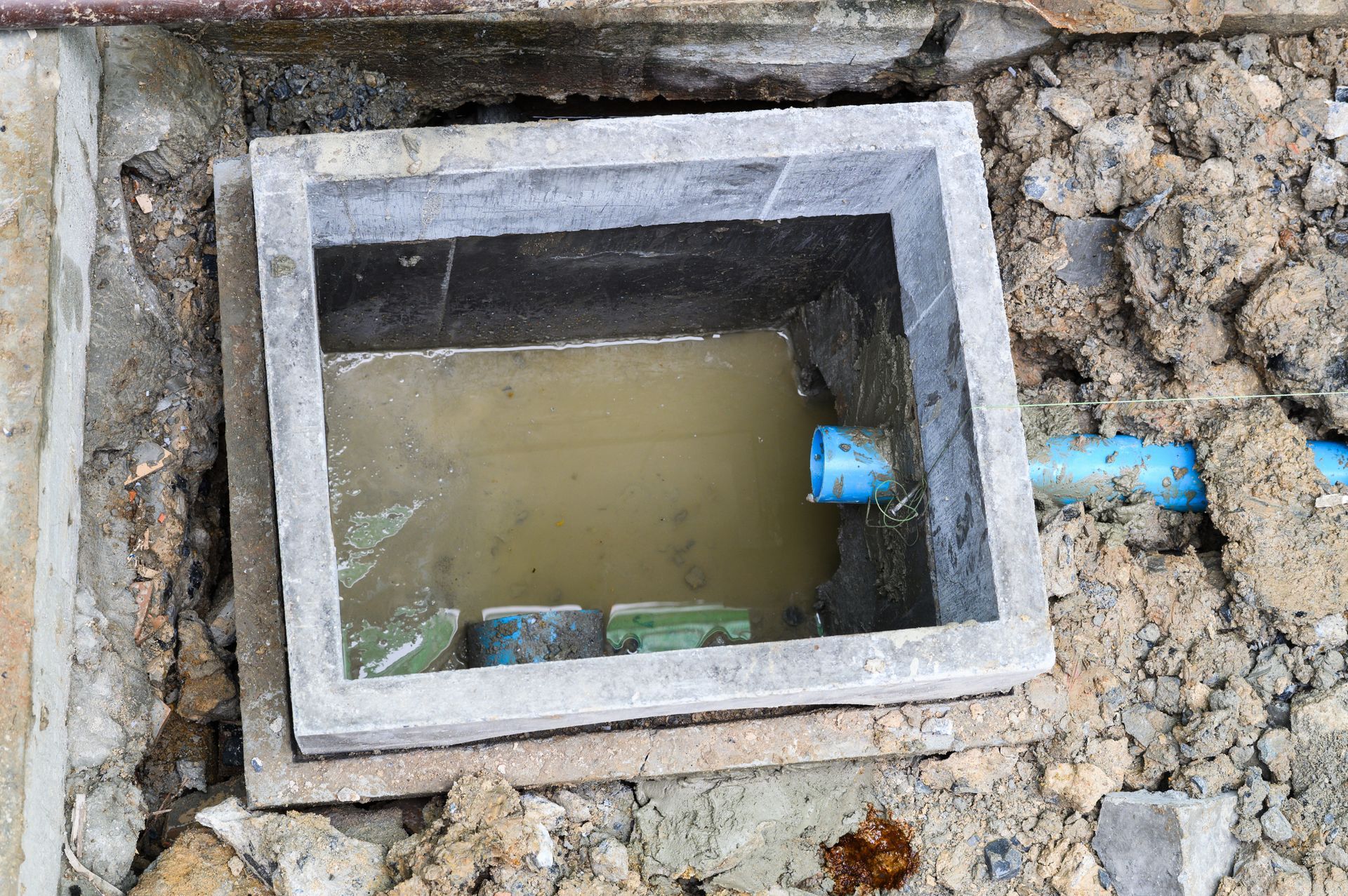 Boîte de drainage en béton ouverte, remplie d'eau trouble et à nu dans la saleté. Tuyau bleu visible.