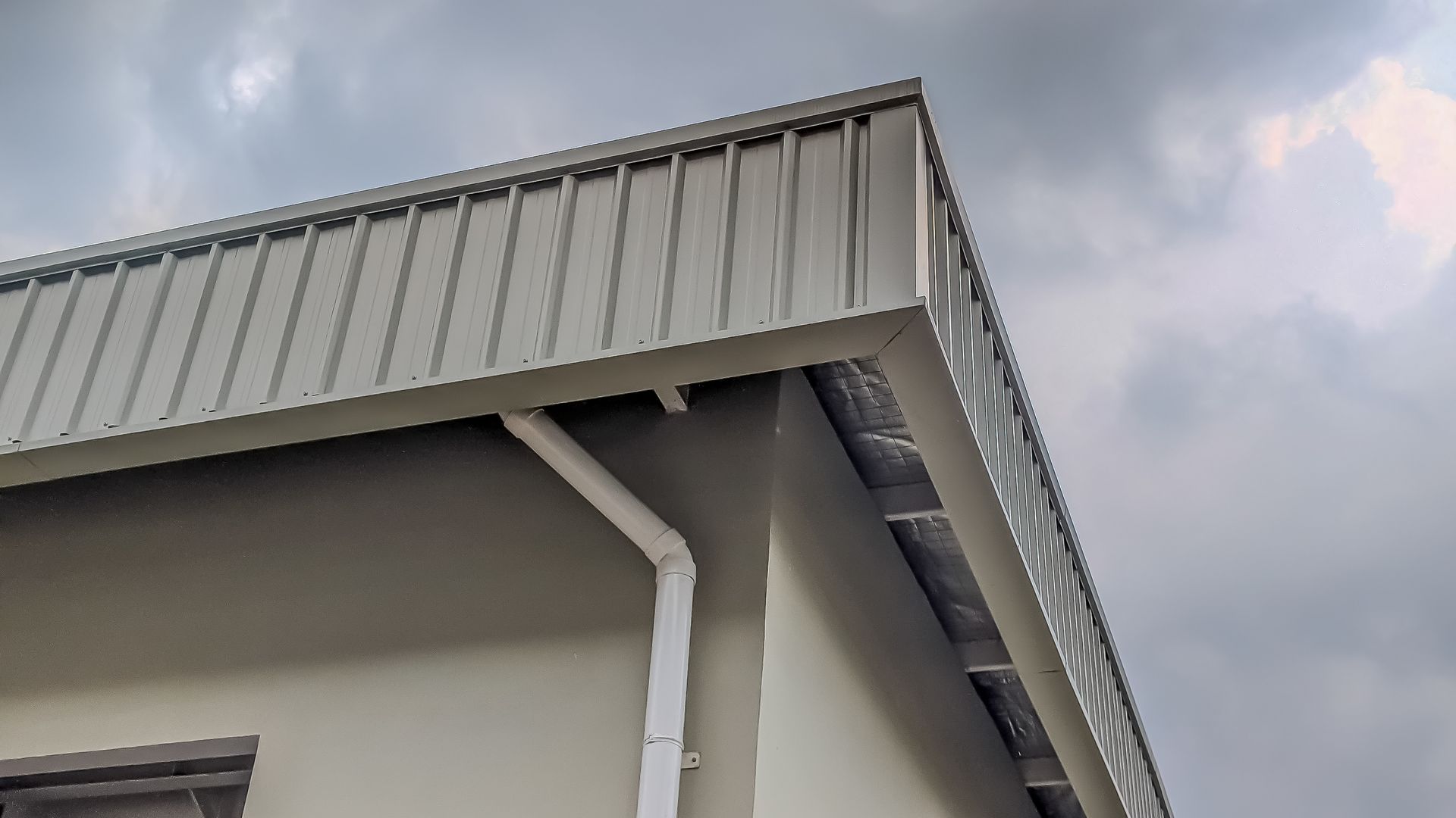 Coin d'un bâtiment avec un toit en métal et une gouttière blanche se détachant sur un ciel nuageux.
