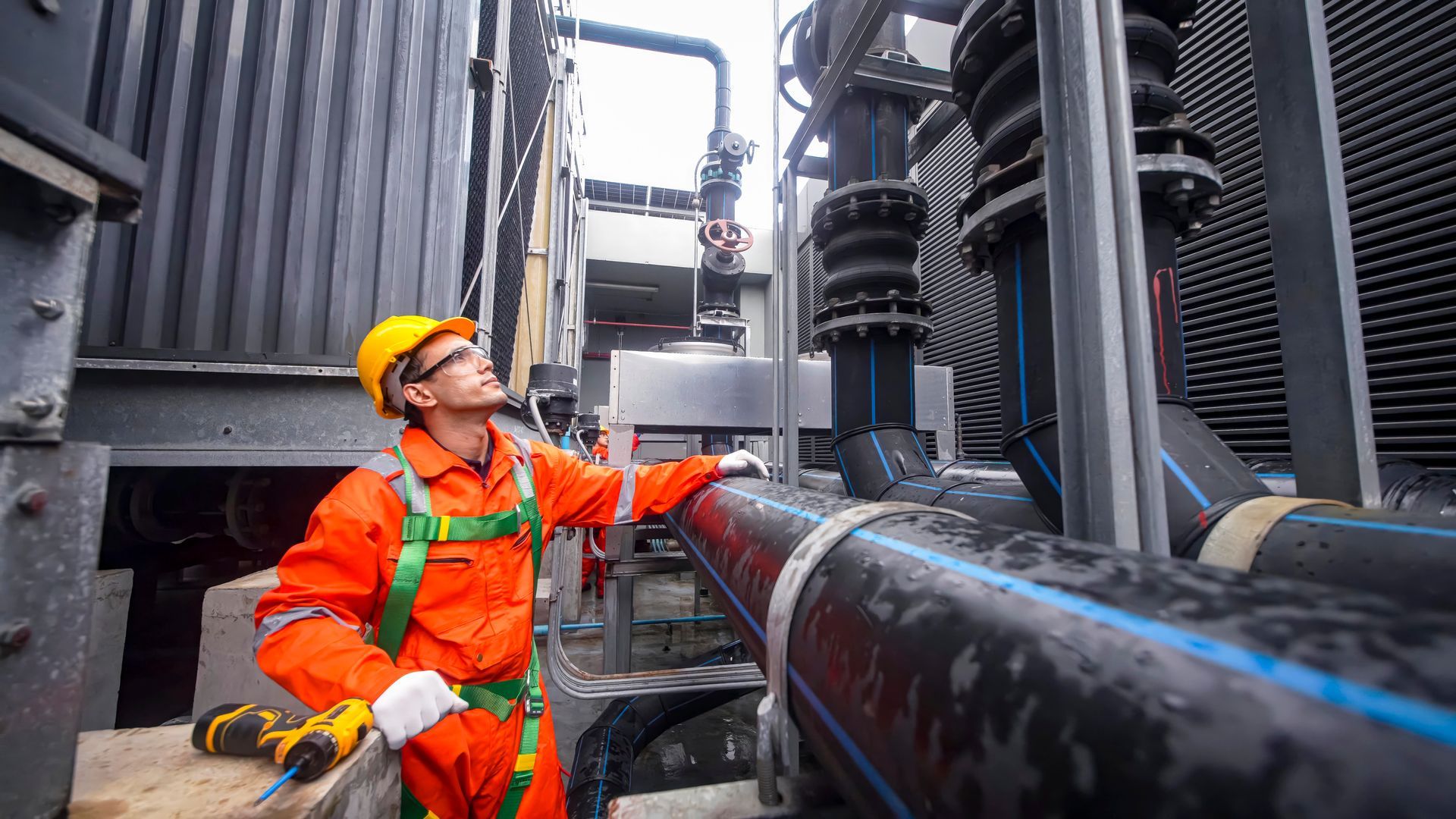 Un ouvrier en combinaison orange et casque de chantier inspecte des canalisations industrielles à l'extérieur.