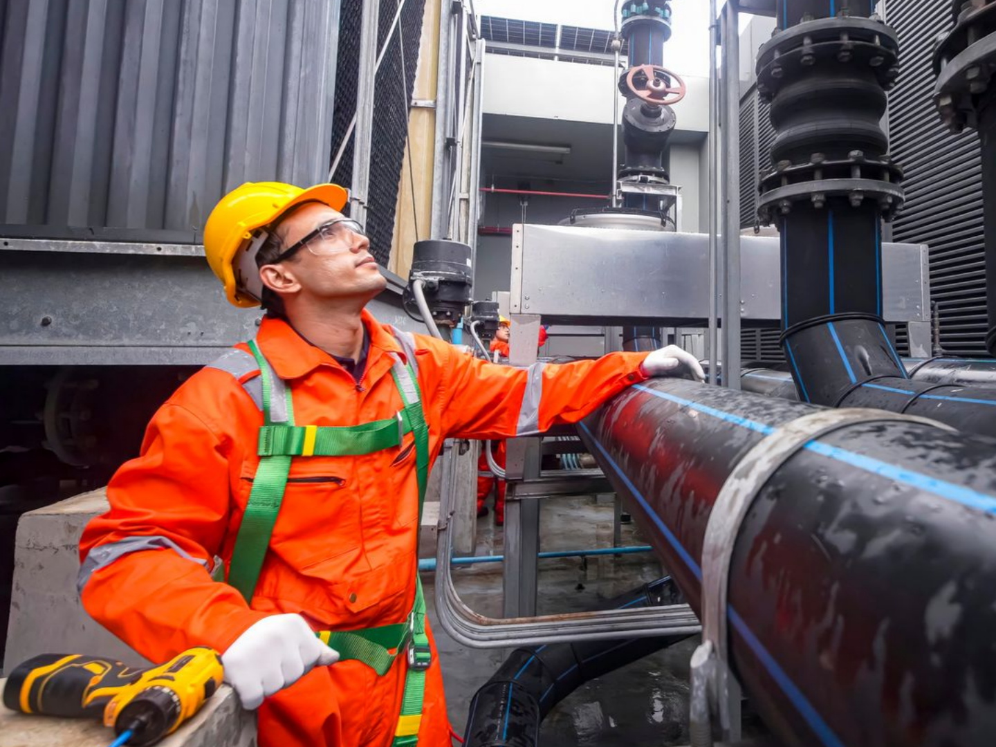 Un ouvrier en combinaison orange et casque de chantier inspecte des canalisations industrielles.