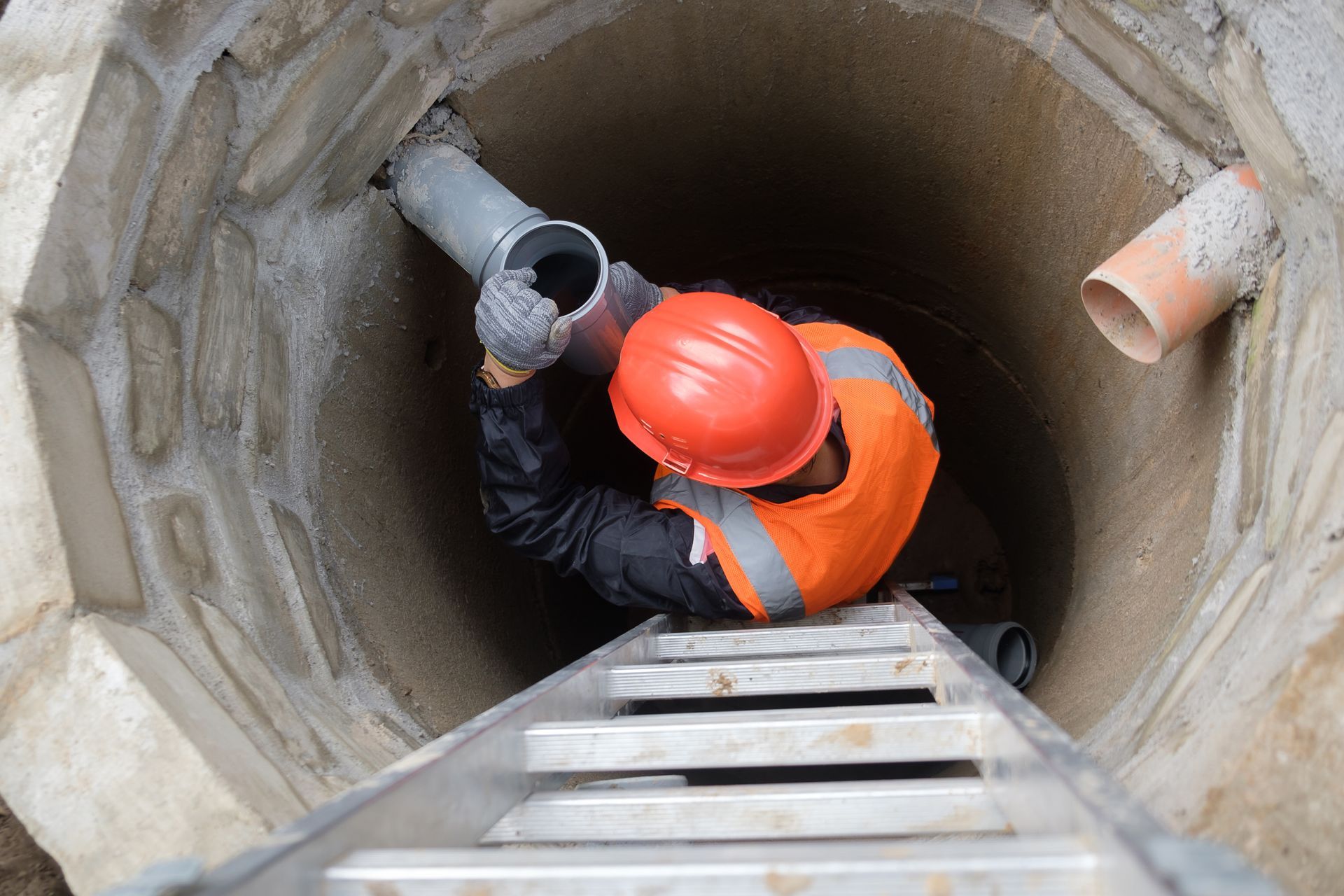 Ouvrier en gilet orange et casque de chantier à l'intérieur d'un puits, en train de raccorder un tuyau.
