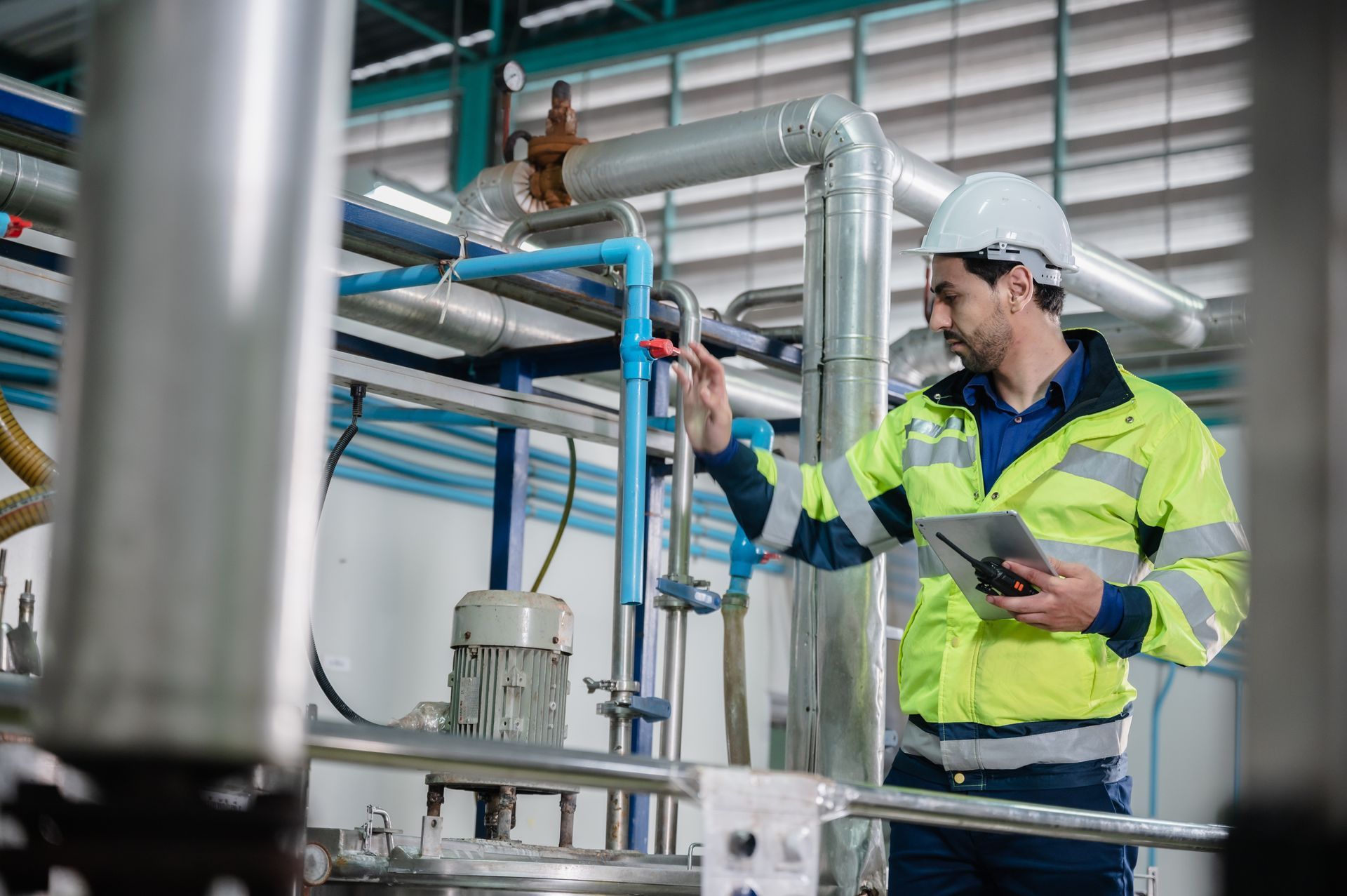 Un homme portant un gilet de sécurité et un casque inspecte des machines dans un environnement industriel, une tablette à la main.