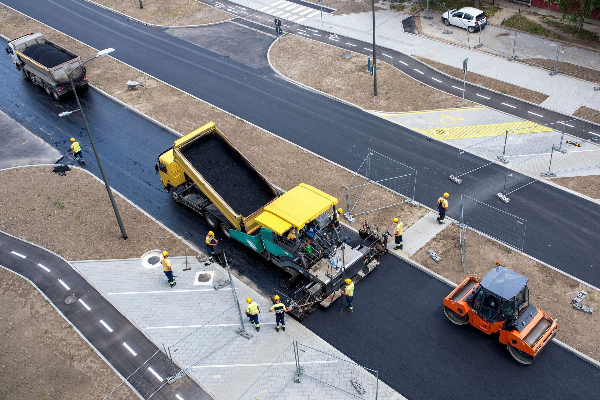 Travaux de pavage : engins jaunes de chantier posant de l’asphalte, avec des ouvriers, des camions et un rouleau compresseur sur une route nouvellement pavée.