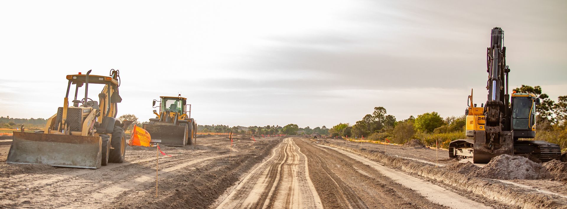 Chantier de construction avec des engins lourds, notamment des bulldozers et une foreuse, sur un chemin de terre sous un ciel nuageux.