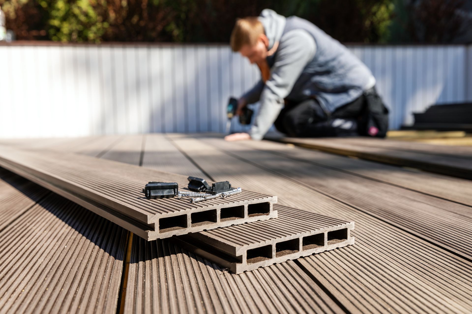 Un homme installe une terrasse composite à l'extérieur à l'aide d'une perceuse ; les lames et la quincaillerie sont visibles au premier plan.