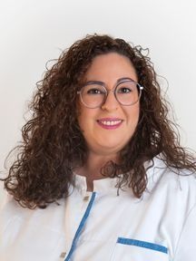Una mujer con cabello rizado y gafas lleva una bata blanca y sonríe.