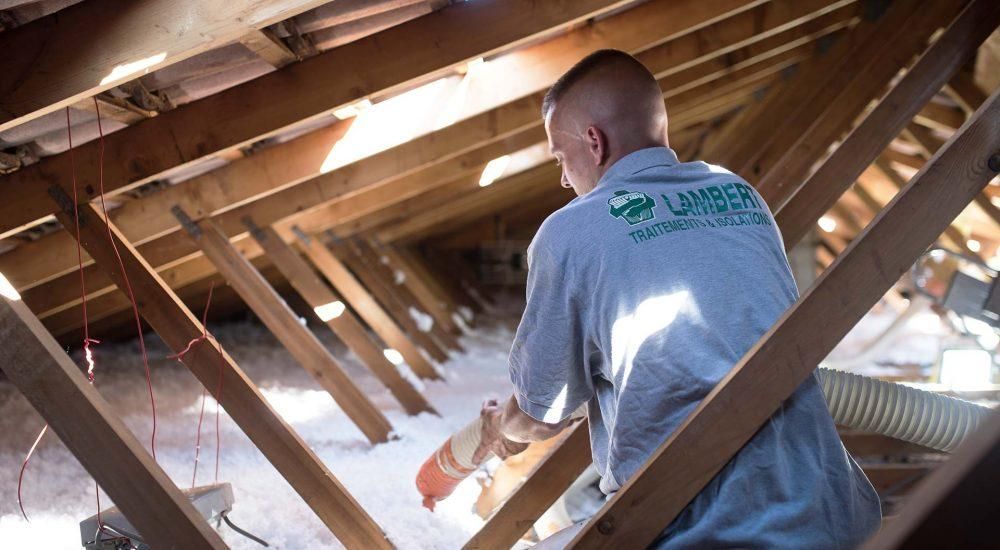 Un homme installe de l'isolant dans un grenier. Poutres en bois, isolant blanc, il porte une chemise grise.