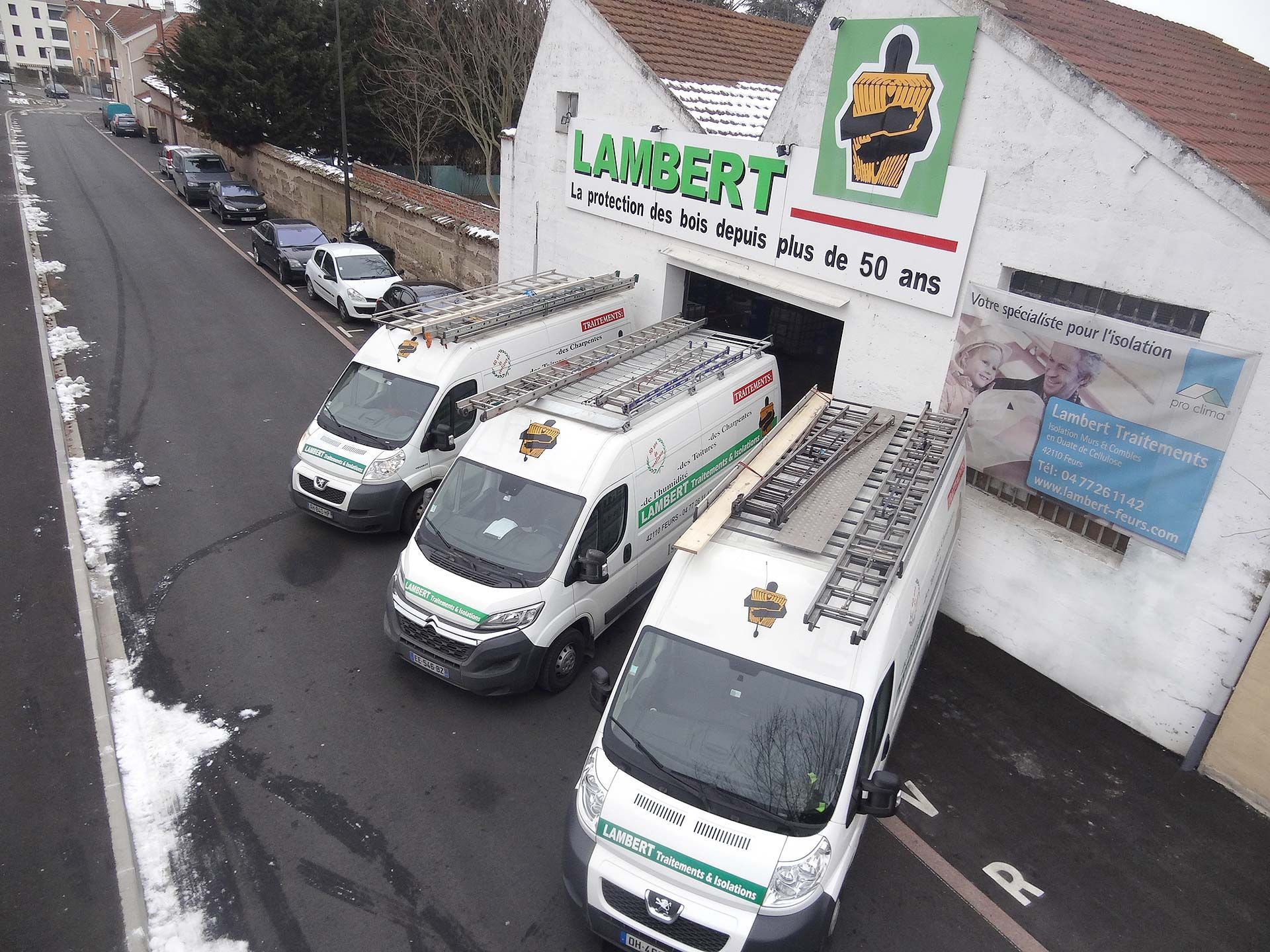 Trois fourgonnettes blanches avec des échelles sont garées devant un bâtiment portant l'inscription « Lambert ». Il y a de la neige au sol.