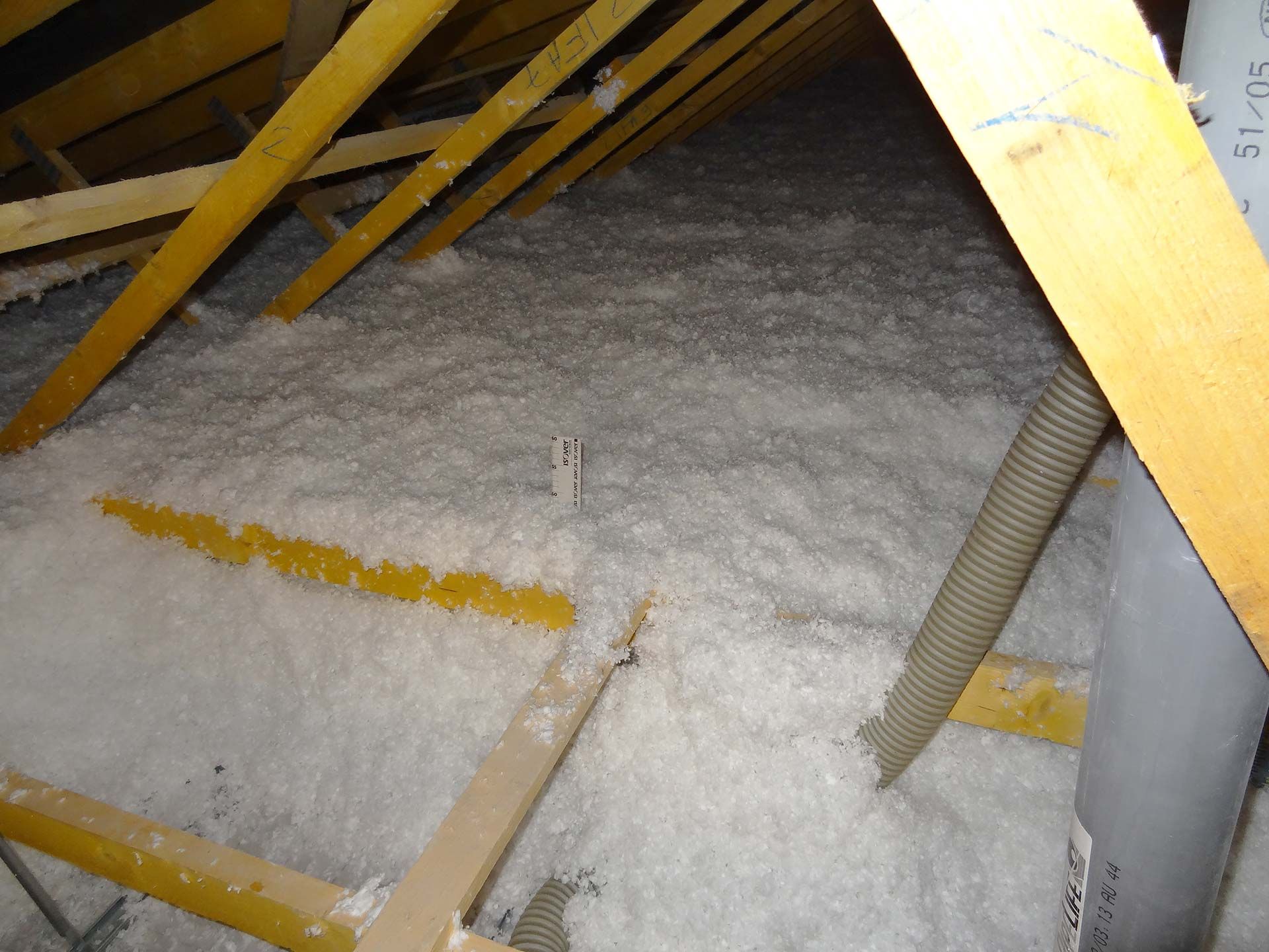 Le grenier est rempli d'isolant blanc entre les chevrons en bois. Un tuyau de ventilation gris est présent.