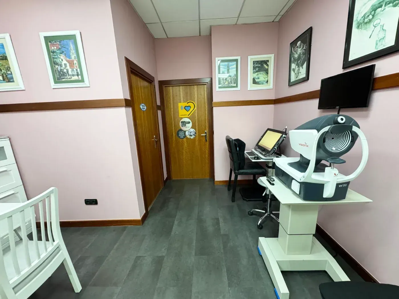 Una sala de examen con equipo ocular, un escritorio, una silla blanca y cuadros en paredes rosas.