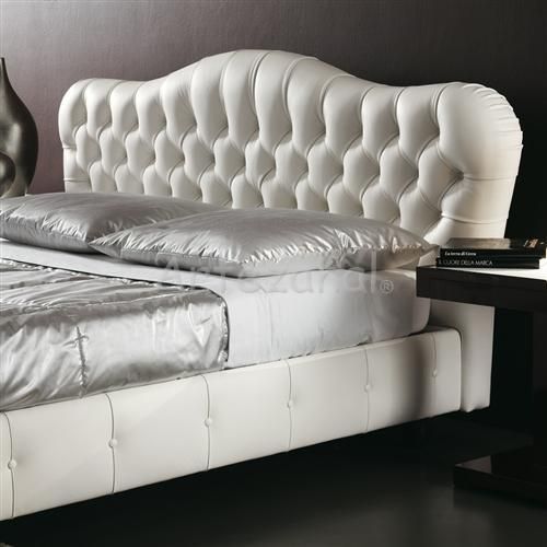 Cama de couro branco com capitonê, roupa de cama prateada e mesa de cabeceira preta.