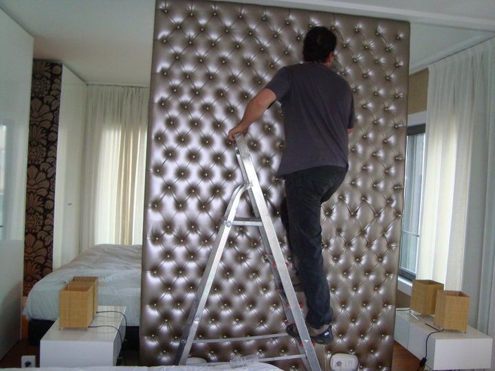 Homem em escada instalando uma cabeceira grande, estofada e prateada em um quarto.