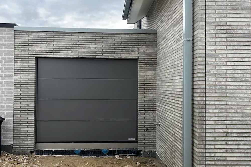 Graues Garagentor in einer hellen Ziegelwand mit einer Dachrinne auf der rechten Seite.