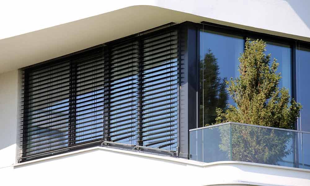 Modernes Gebäude mit schwarzen verstellbaren Außenjalousien und einem Glasbalkon mit einem kleinen Baum.