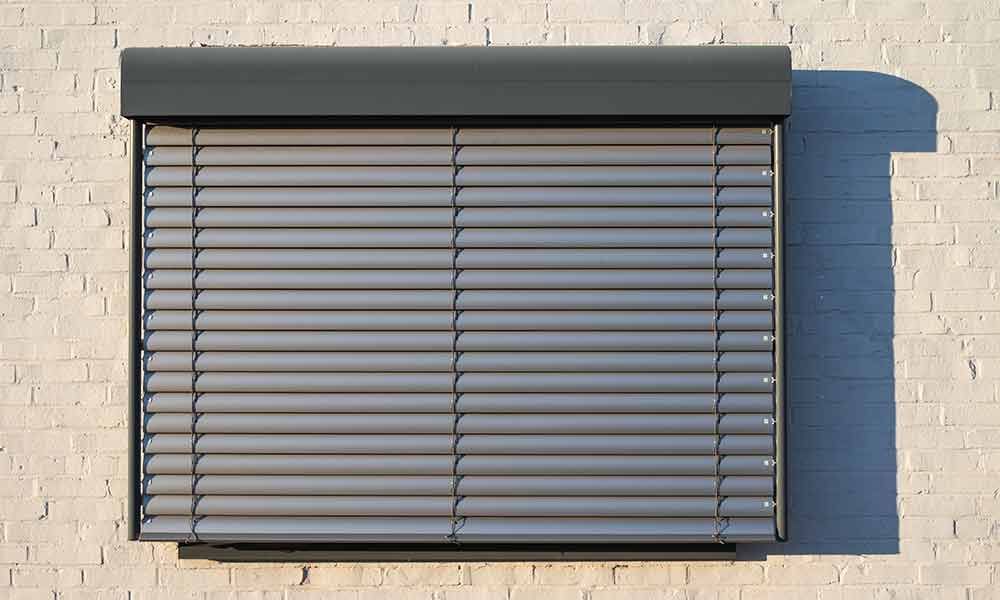 Grauer Rollladen an einer weißen Ziegelwand, der ein Fenster teilweise verdeckt.