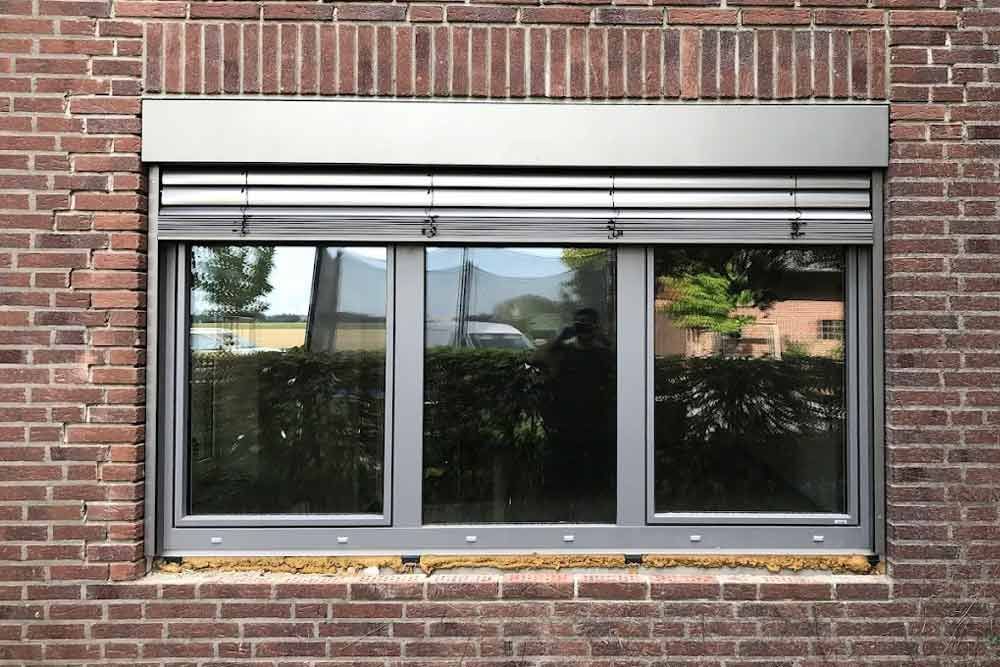 Fenster mit geschlossenen silbernen Jalousien und einem dunklen Rahmen in einer Ziegelwand.