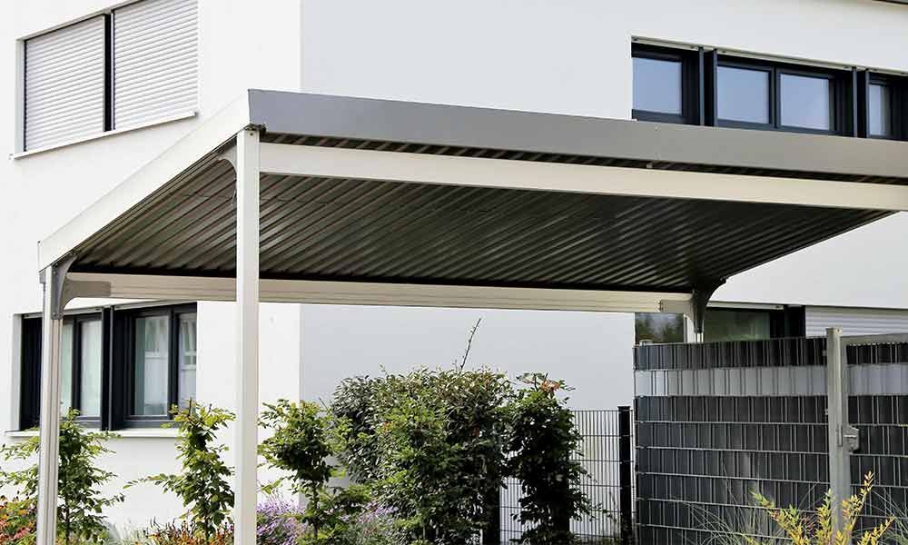 Carport mit grauem Dach, weißem Rahmen und grünen Sträuchern vor einem weißen Gebäude.