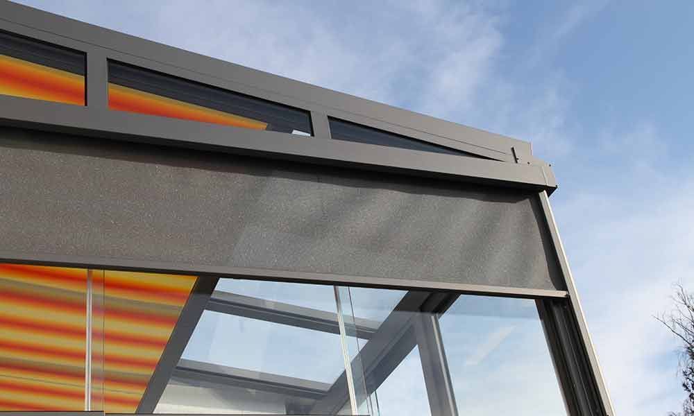 Dunkelgrauer Außenschirm teilweise über einer Glasstruktur. Orange und gelbe Akzente sichtbar. Blauer Himmel.