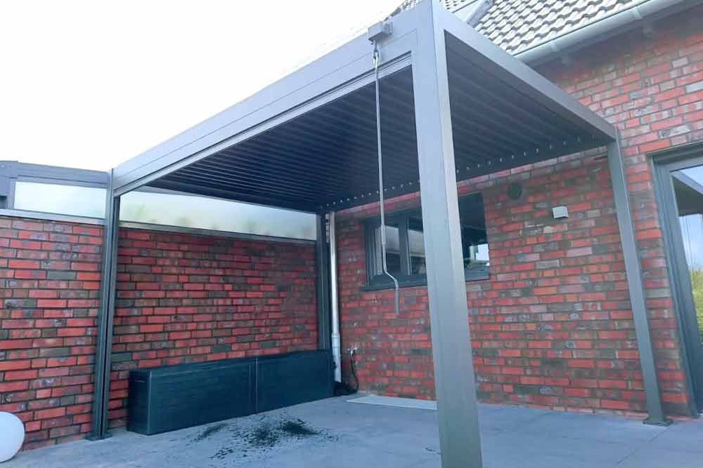 An einem Backsteingebäude befestigte Pergolakonstruktion; anthrazitfarbener Rahmen mit horizontalen Lamellen.