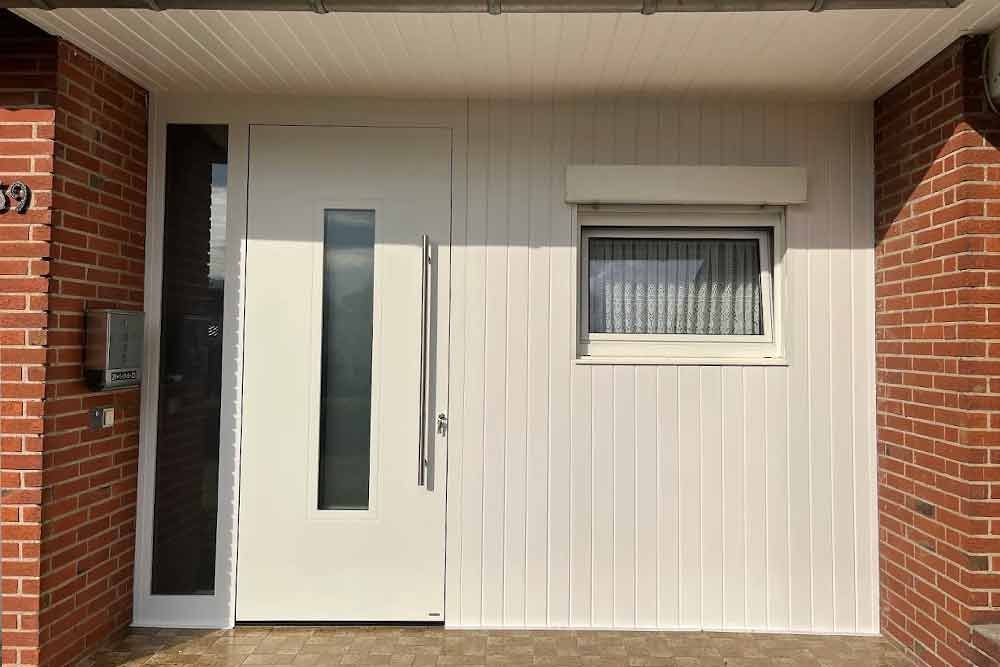 Weiße Haustür mit Glasscheibe und Seitenlicht neben einem Backsteingebäude.