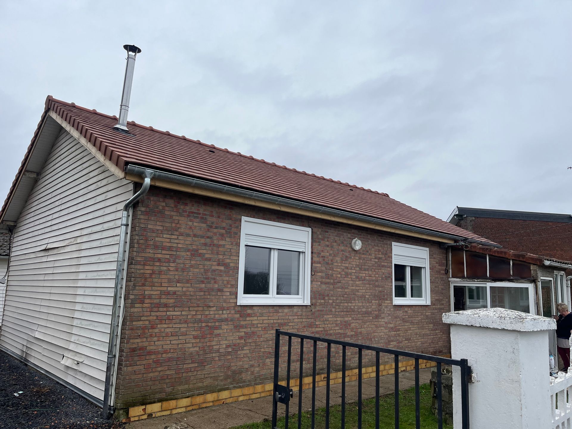 Maison en briques au toit de tuiles rouges, aux fenêtres blanches et à la clôture noire, sous un ciel nuageux.