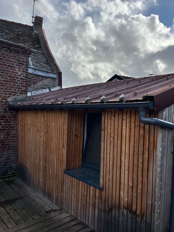 Abri rustique aux parois de bambou et au toit de tuiles, jouxtant un bâtiment en briques sous un ciel nuageux.