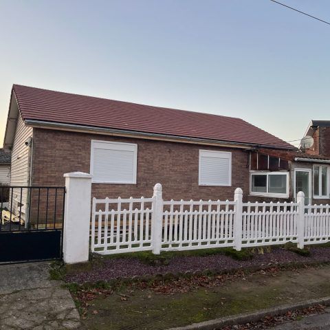 Maison de plain-pied en briques, avec toit rouge, volets blancs et clôture blanche en bordure de rue.