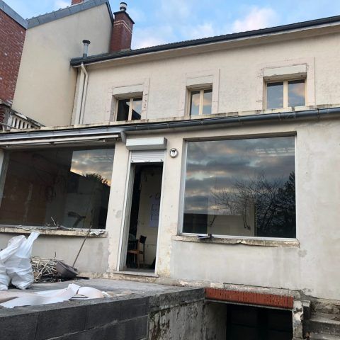 Extérieur d'une maison à deux étages avec porte ouverte, grandes fenêtres et patio partiellement aménagé.