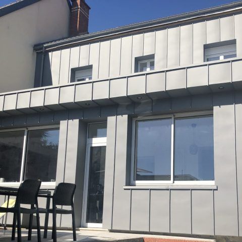 Maison moderne grise avec de grandes fenêtres, une terrasse et deux chaises noires sous un auvent.