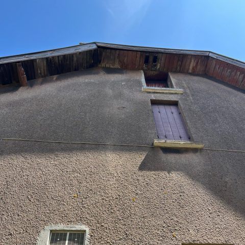Vue en contre-plongée d'un bâtiment en stuc gris avec une petite fenêtre à volets sous la toiture et un ciel bleu au-dessus.