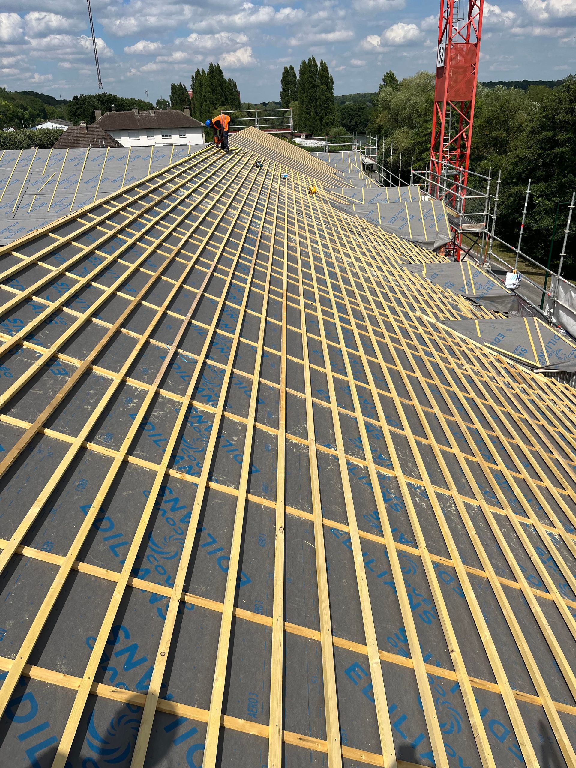 Toiture en construction avec des liteaux jaunes, des ouvriers et une grue rouge sous un ciel bleu