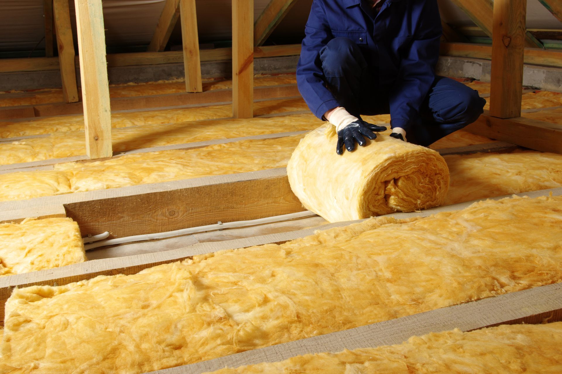 Une personne vêtue d’une tenue de travail bleue installe de l’isolant en fibre de verre jaune dans les combles.