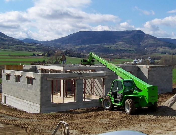 Un chariot télescopique vert soulève des matériaux sur un bâtiment en bloc de béton en construction avec des montagnes en arrière-plan.