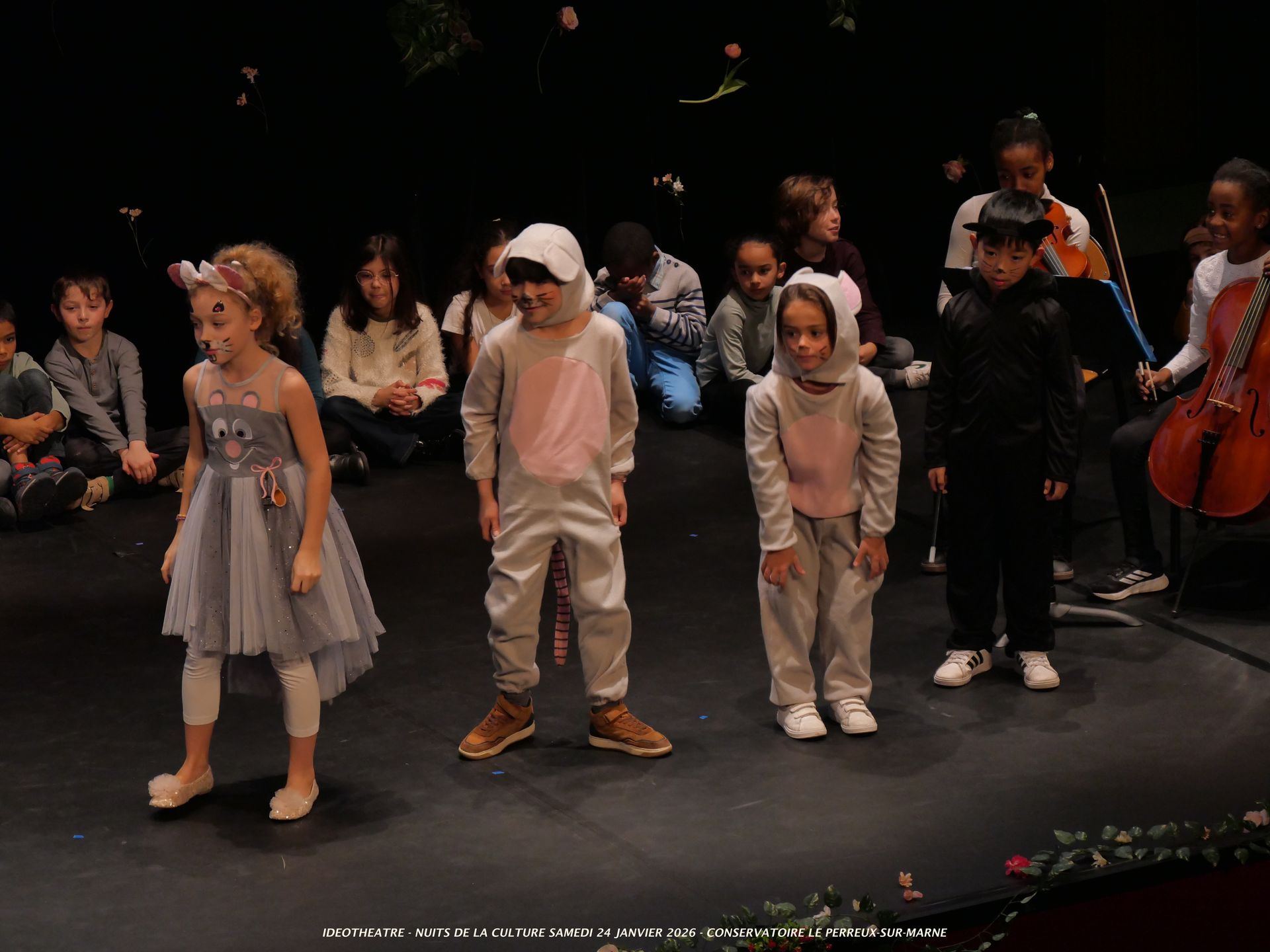 Des enfants costumés sur scène. L'un porte un costume de souris, une autre une robe avec un nœud. D'autres les regardent.