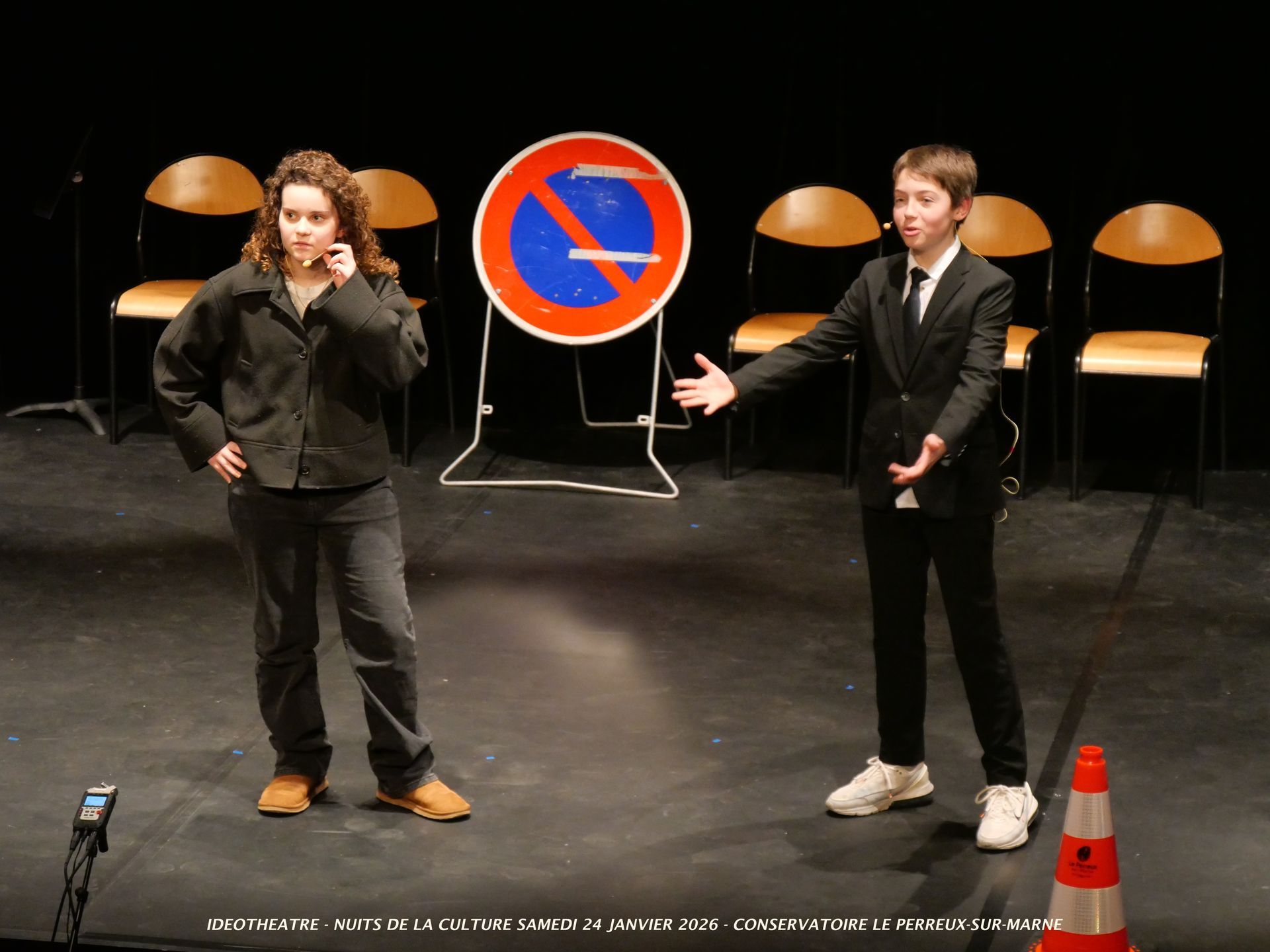 Deux personnes sur scène avec un panneau d'interdiction de stationner, l'une faisant des gestes, l'autre au téléphone.
