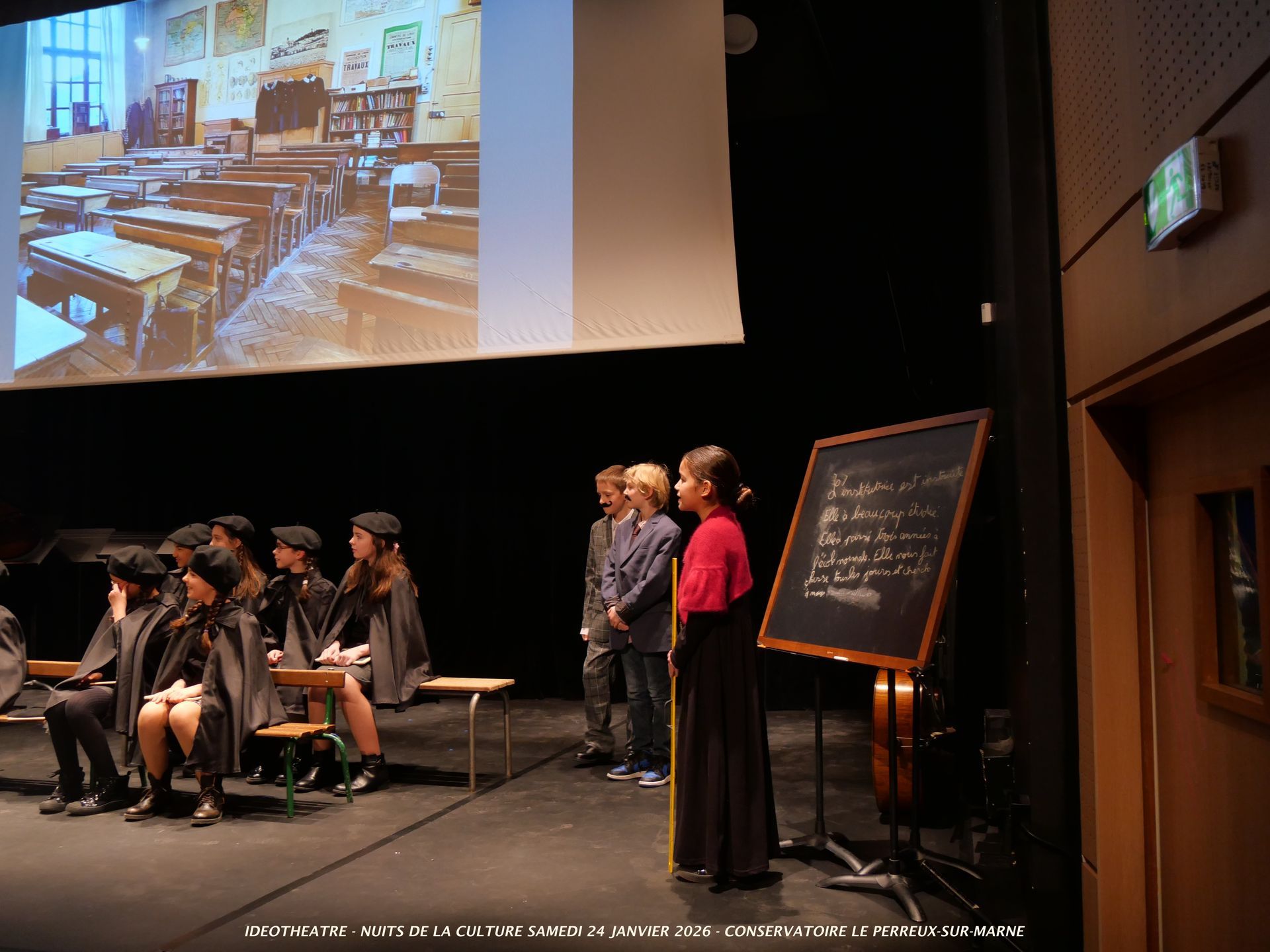 Des élèves sur scène, certains en capes, devant un tableau noir et l'image projetée d'une salle de classe.