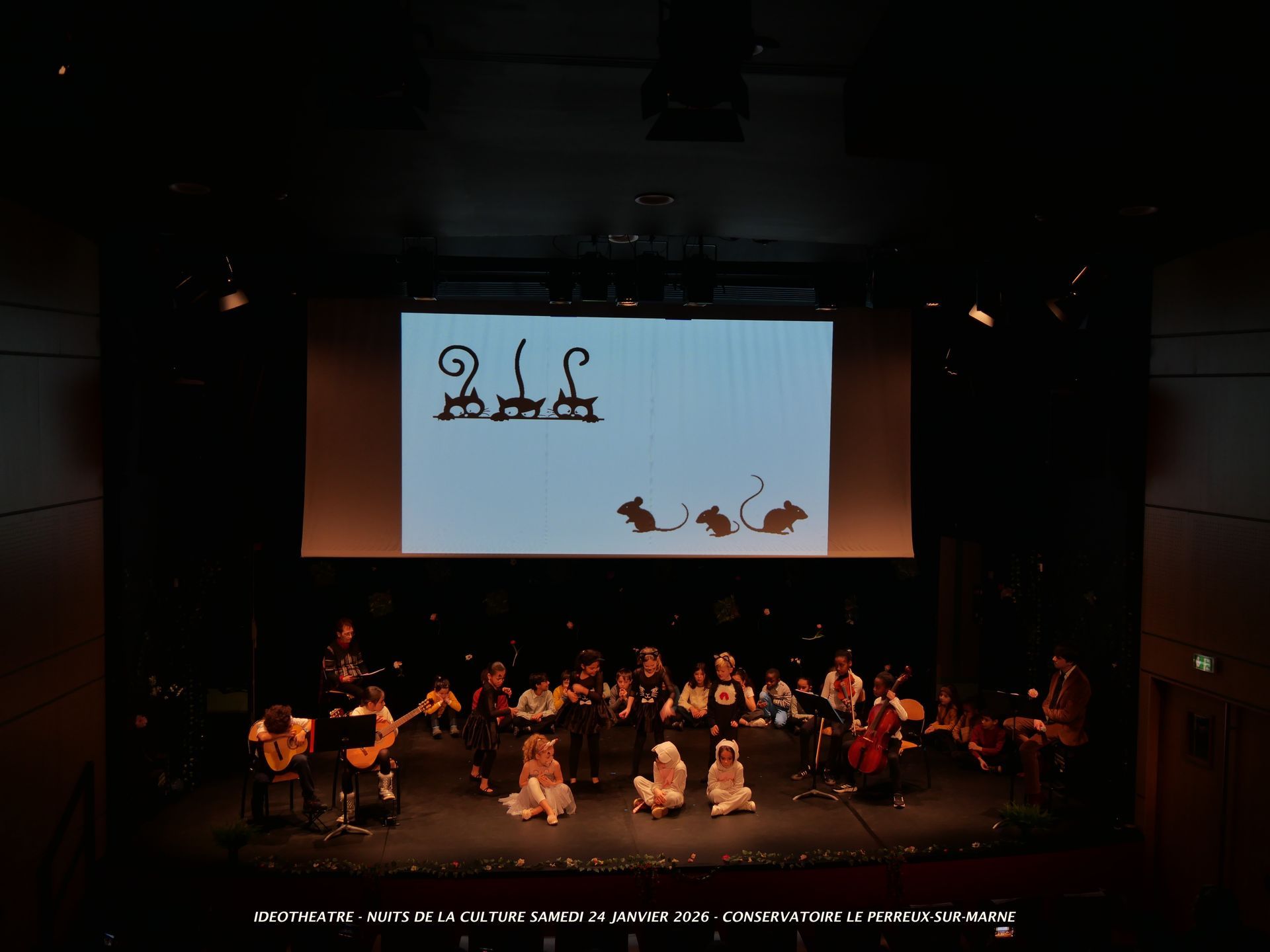 Scène de théâtre avec des musiciens et des artistes sous un écran diffusant des silhouettes animées de singes et de souris.