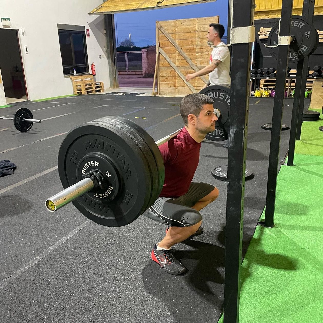 Hombre con camisa marrón haciendo sentadillas con una barra pesada en el gimnasio.