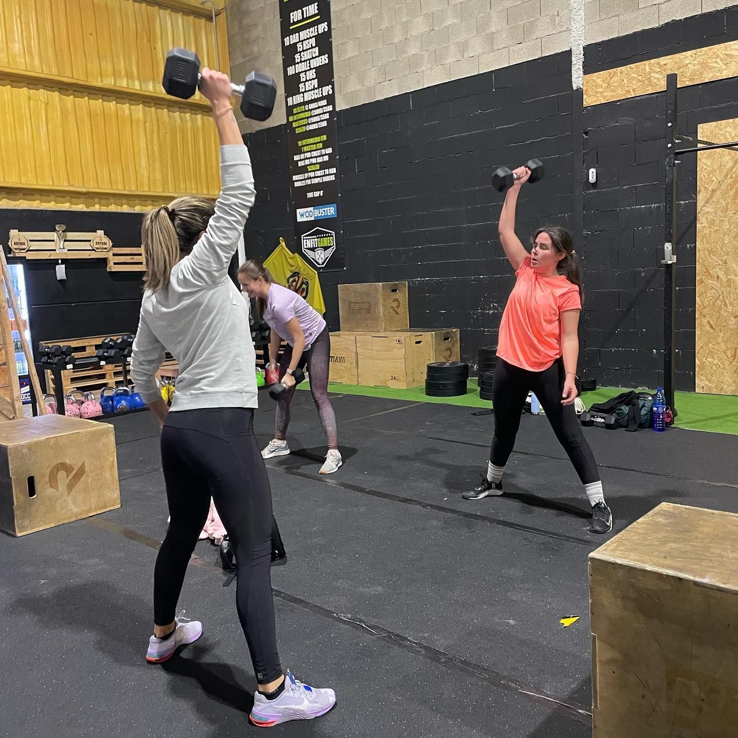Tres mujeres levantan mancuernas en un gimnasio. Una lleva una pesa por encima de la cabeza, las otras las sostienen a la altura de los hombros.