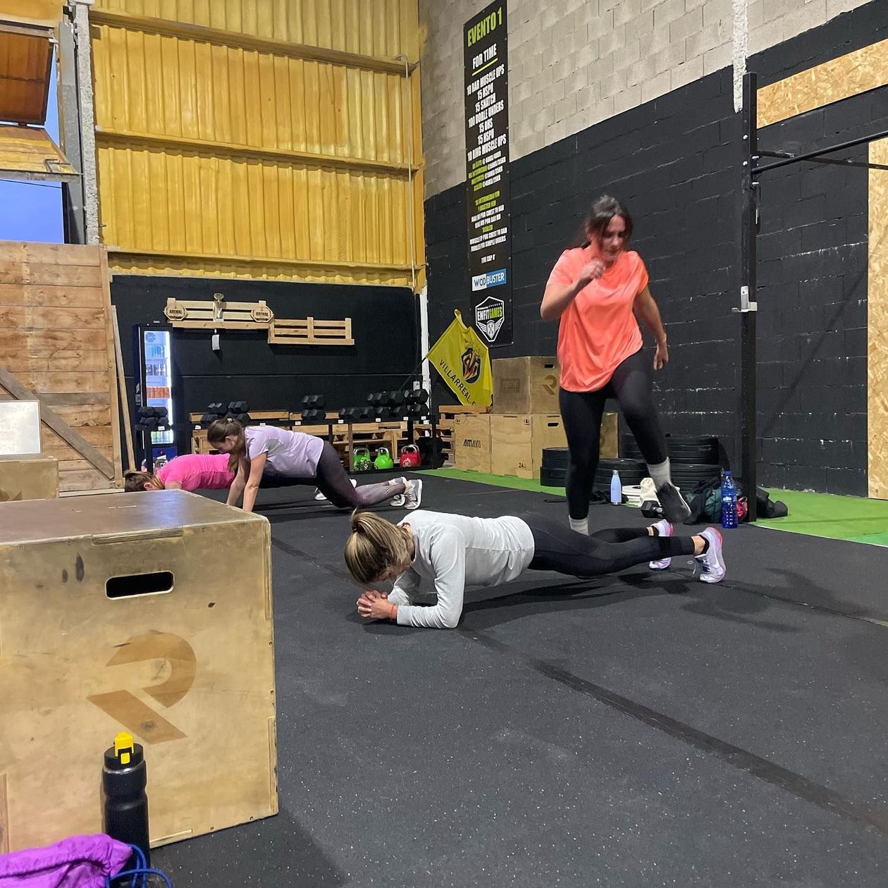 Tres mujeres haciendo ejercicio en un gimnasio: planchas, flexiones y saltos. Suelo negro, techo amarillo.