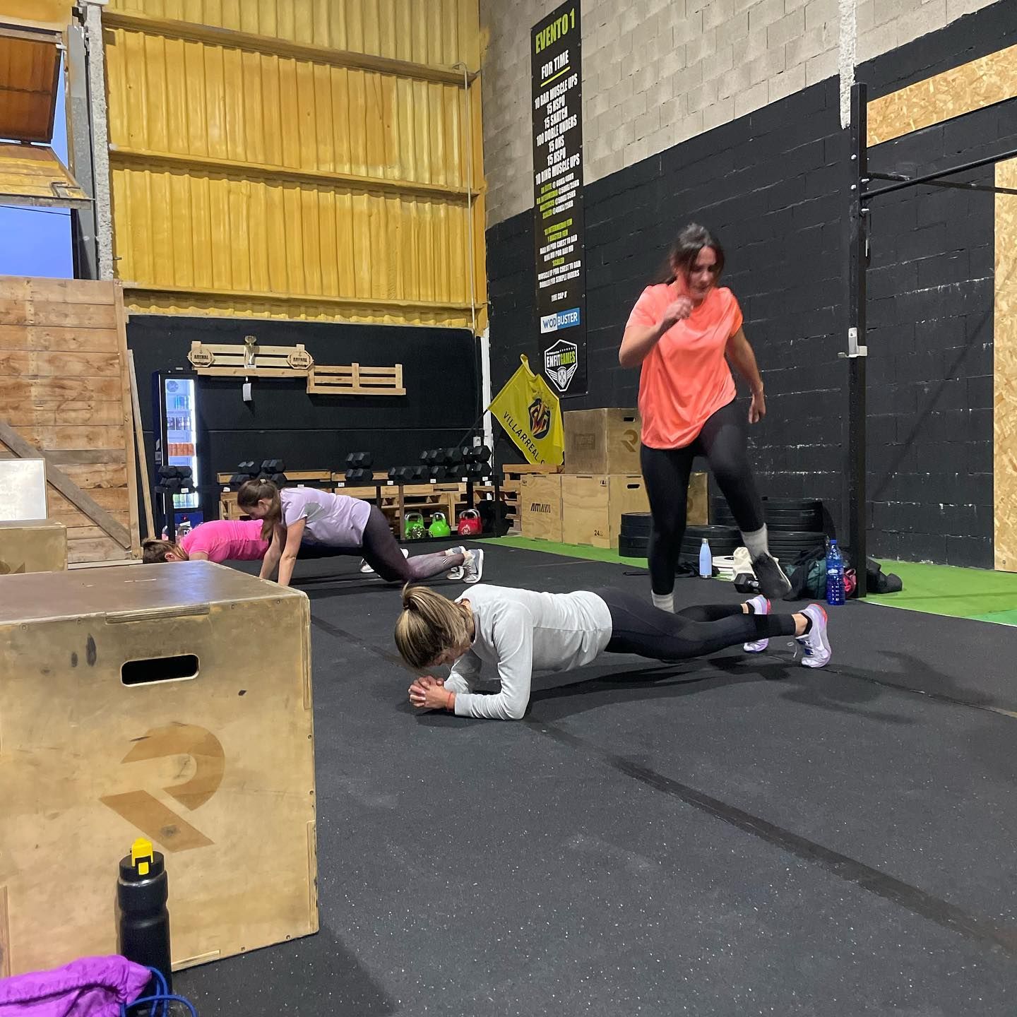 Tres mujeres haciendo ejercicio en un gimnasio: planchas, flexiones y saltos. Suelo negro, techo amarillo.