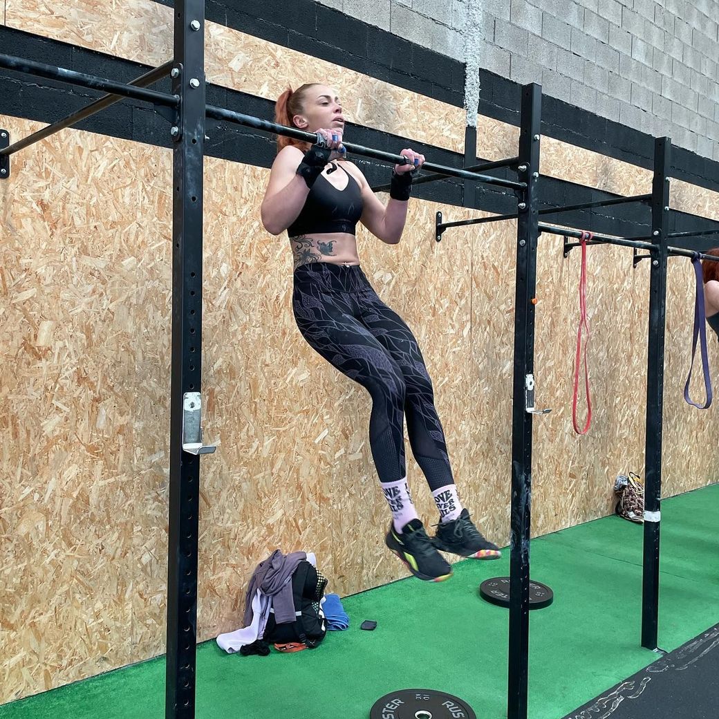 Mujer haciendo una dominada en un gimnasio, vestida con ropa deportiva negra, con un tatuaje en el abdomen.