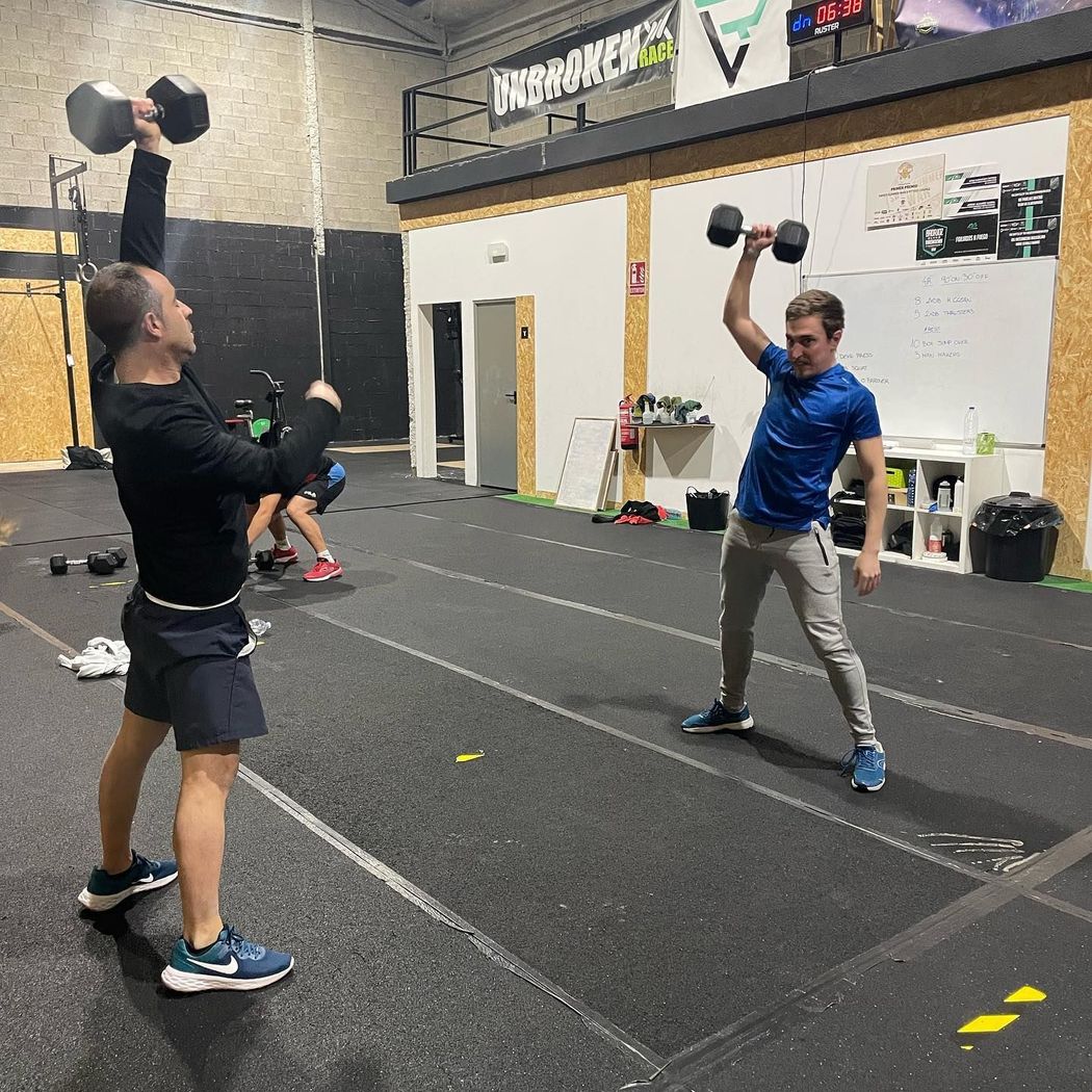 Dos hombres en un gimnasio levantando mancuernas. Interior: suelo negro, paredes blancas.