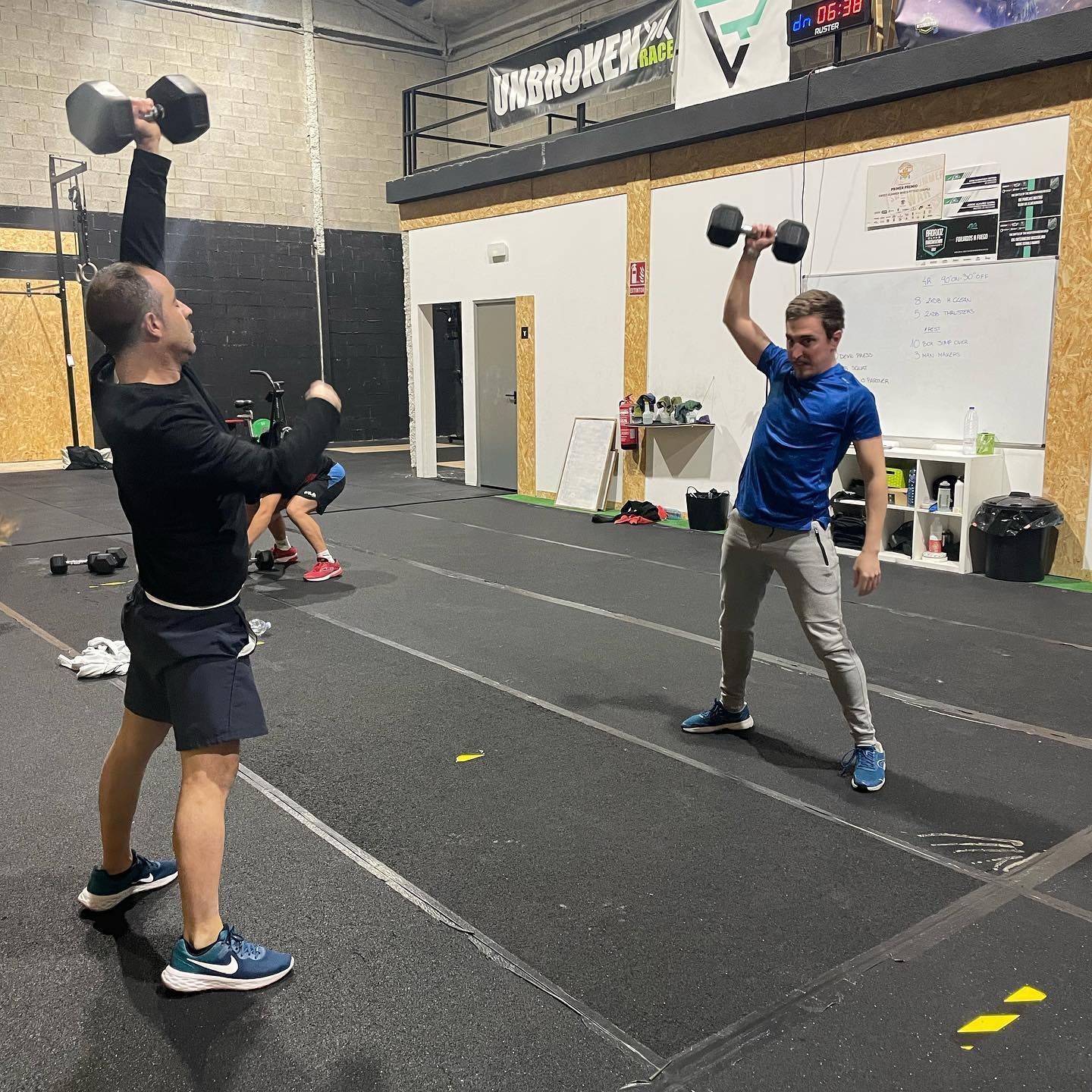 Dos hombres en un gimnasio levantando mancuernas. Interior: suelo negro, paredes blancas.