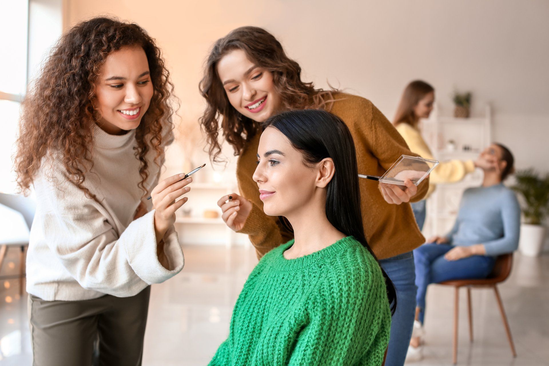 Deux coiffeuses travaillent sur une cliente assise dans un salon lumineux, l'une lui appliquant du maquillage et l'autre la coiffant.