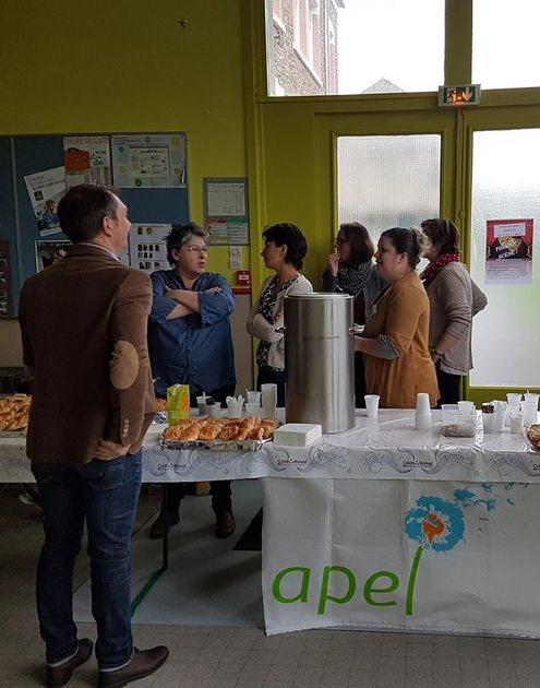 Des personnes étaient rassemblées autour d'une table de rafraîchissements lors d'un événement en intérieur, avec une banderole « apel ».