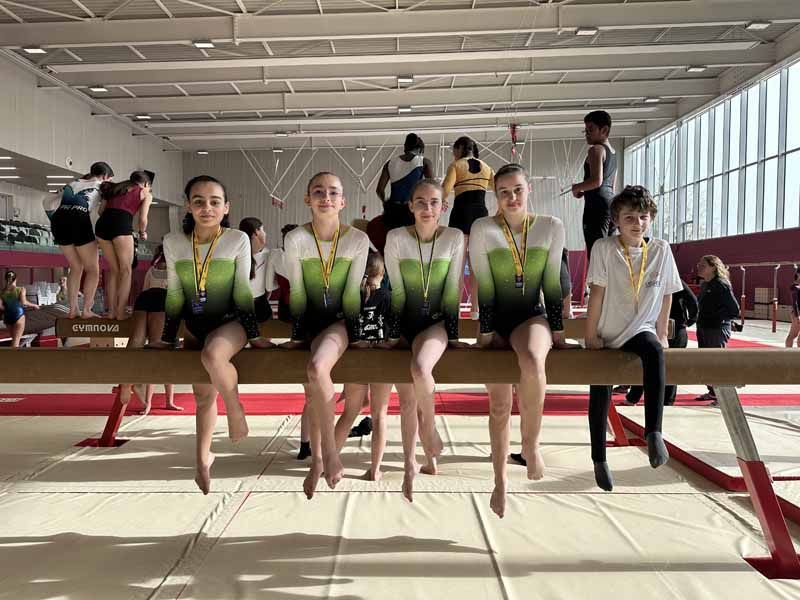 Cinq gymnastes en justaucorps verts et noirs sont assises sur une poutre d'équilibre dans un gymnase lumineux, attendant entre deux exercices.