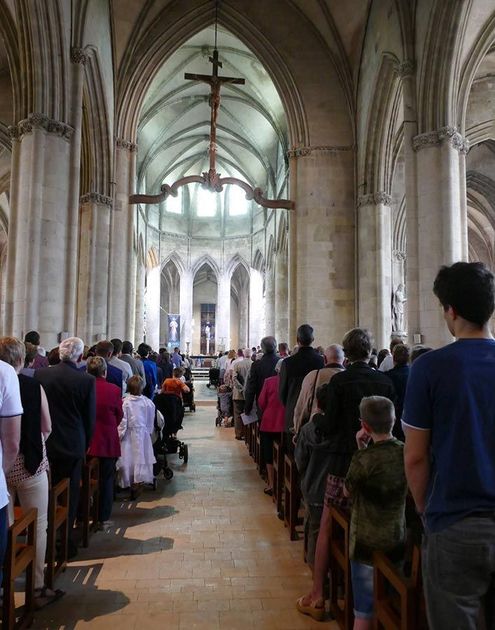 Allée bondée d'une cathédrale, avec des fidèles debout sur les bancs sous des voûtes et un crucifix suspendu.